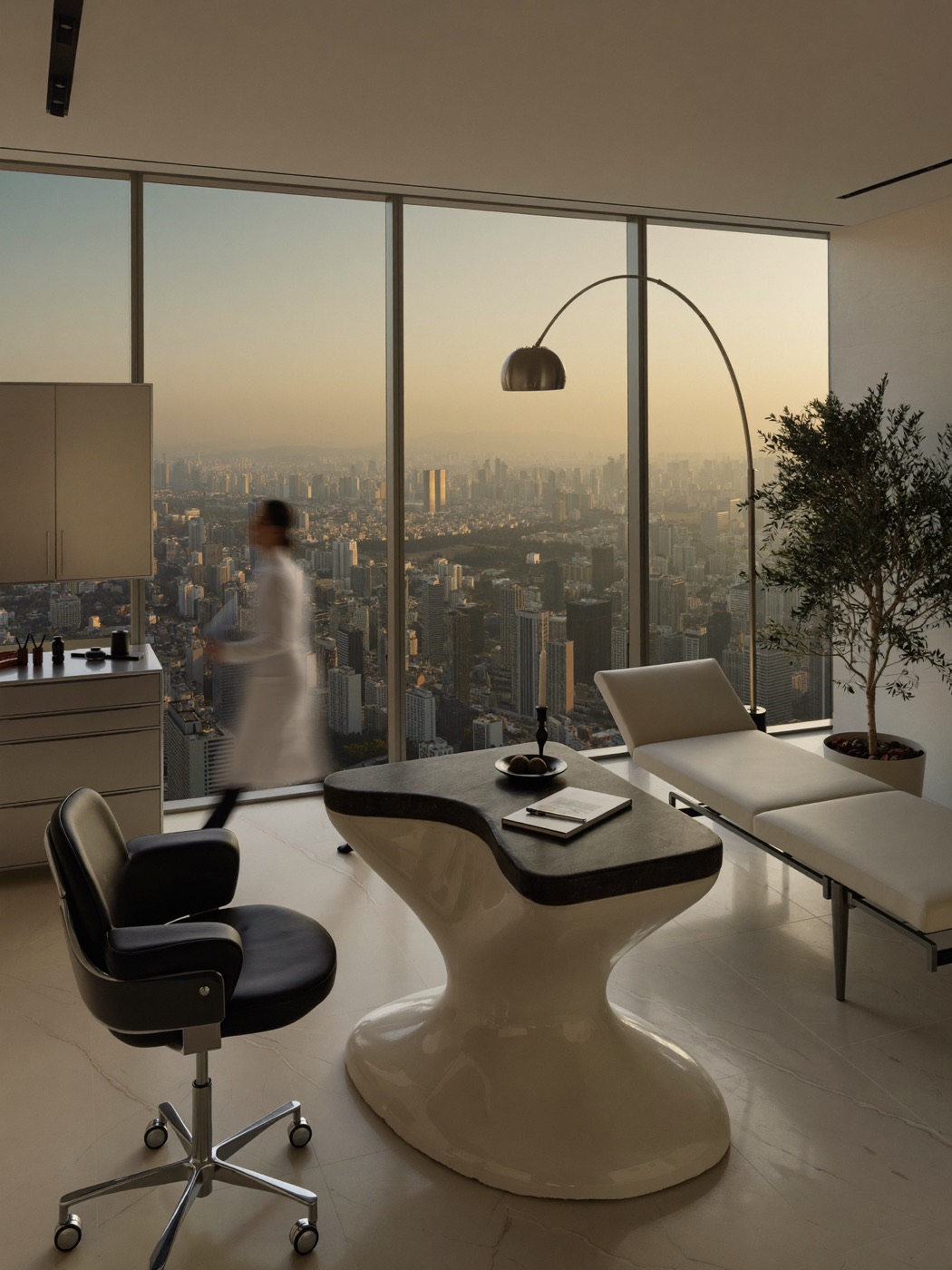 Consultório de alto padrão com mobiliário de design e janela ampla sobre São Paulo ao entardecer; um médico atravessa a sala em borrão de movimento